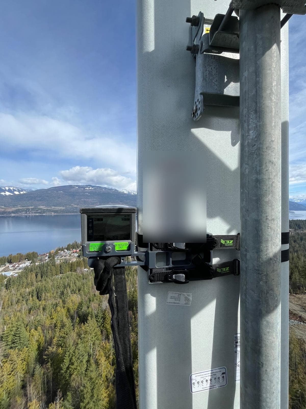 Antenna alignment tool strapped to a cellular sector antenna on a tower, with a lake and snow-capped mountains below