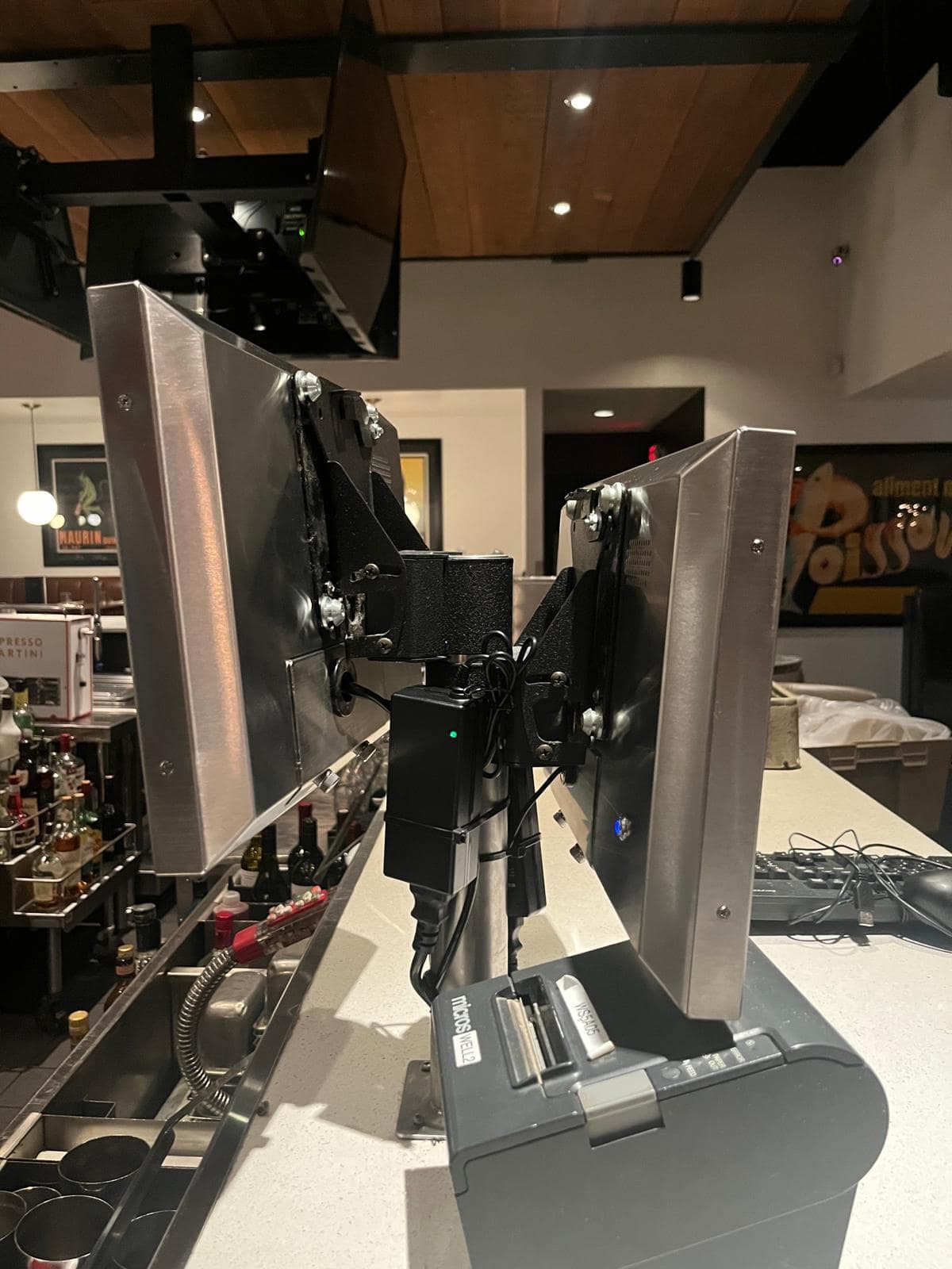 Two stainless-clad point-of-sale terminals on articulated arms above a bar counter, with a receipt printer below