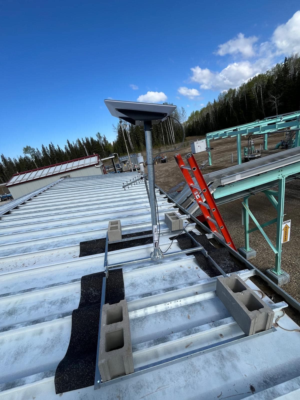 Starlink dish on a tall pole mount weighted with concrete blocks on a standing-seam metal roof, with a directional Yagi antenna alongside and an industrial facility beyond