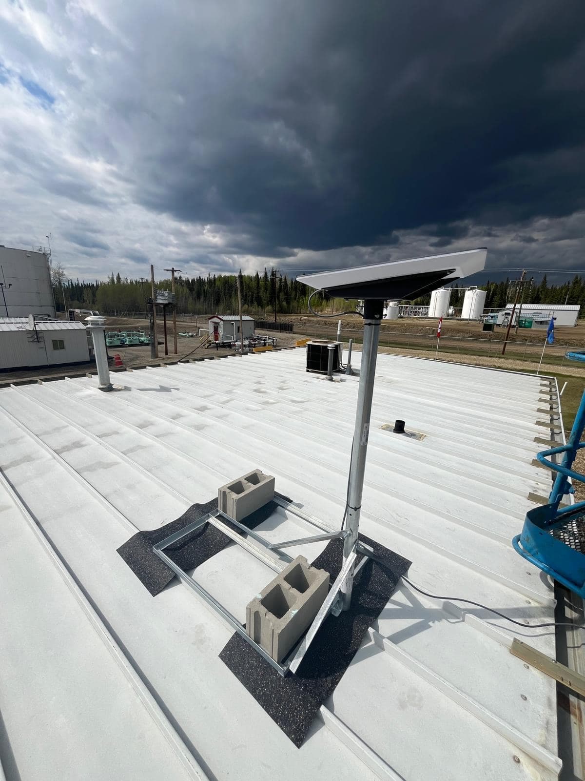 Starlink dish on a galvanised pole with block ballast on a white metal roof, storm clouds overhead and process equipment in the background