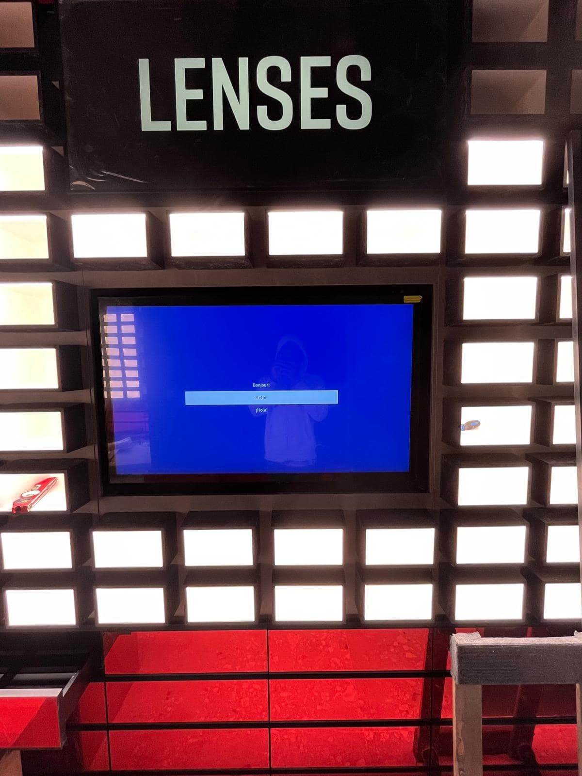 Wall-mounted display screen recessed into a backlit retail merchandising wall beneath a Lenses sign
