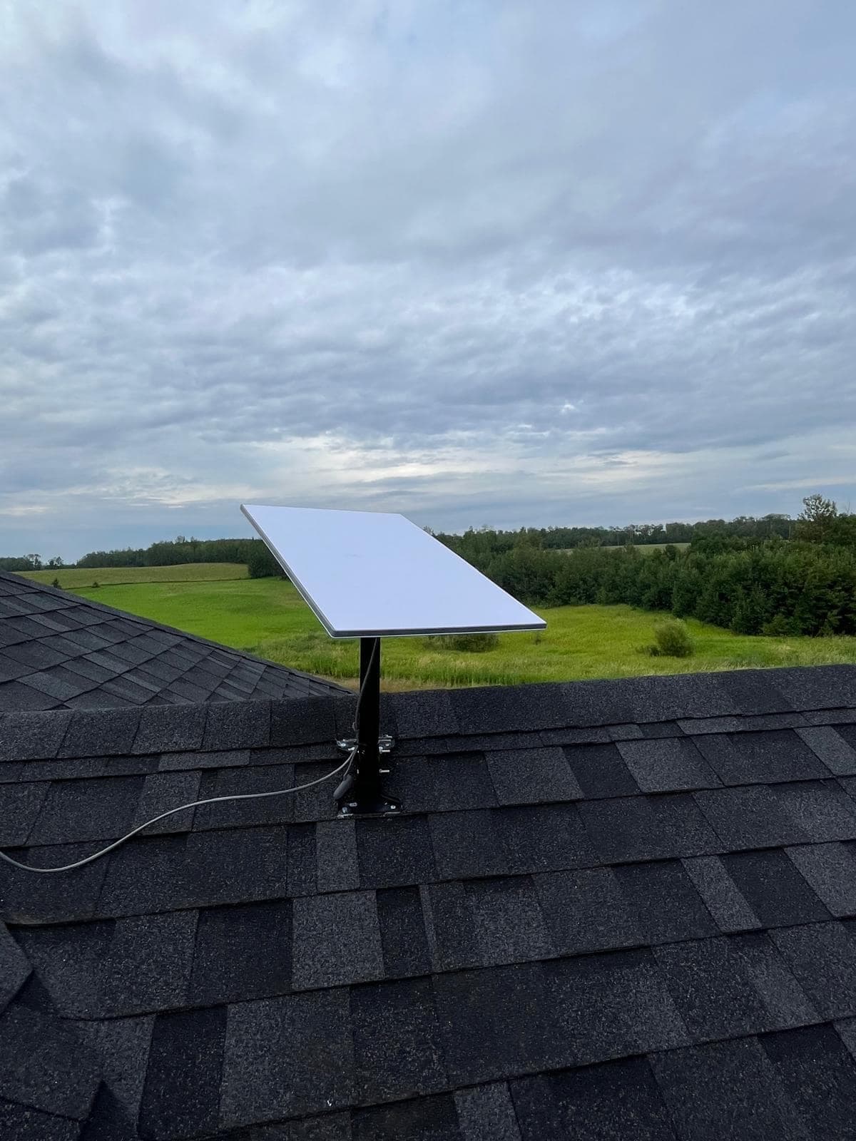 Starlink dish angled toward open sky above a roof ridge, with green fields and bush in the distance